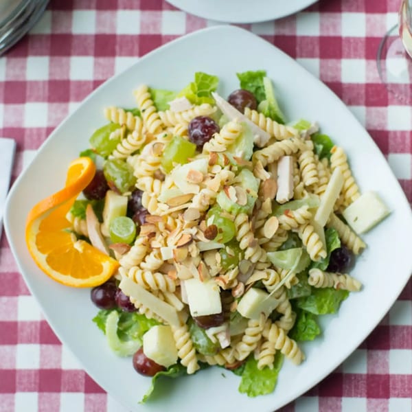 Pasta salad with grapes, cheese, almonds, and an orange slice on a white plate over a red and white checkered tablecloth.