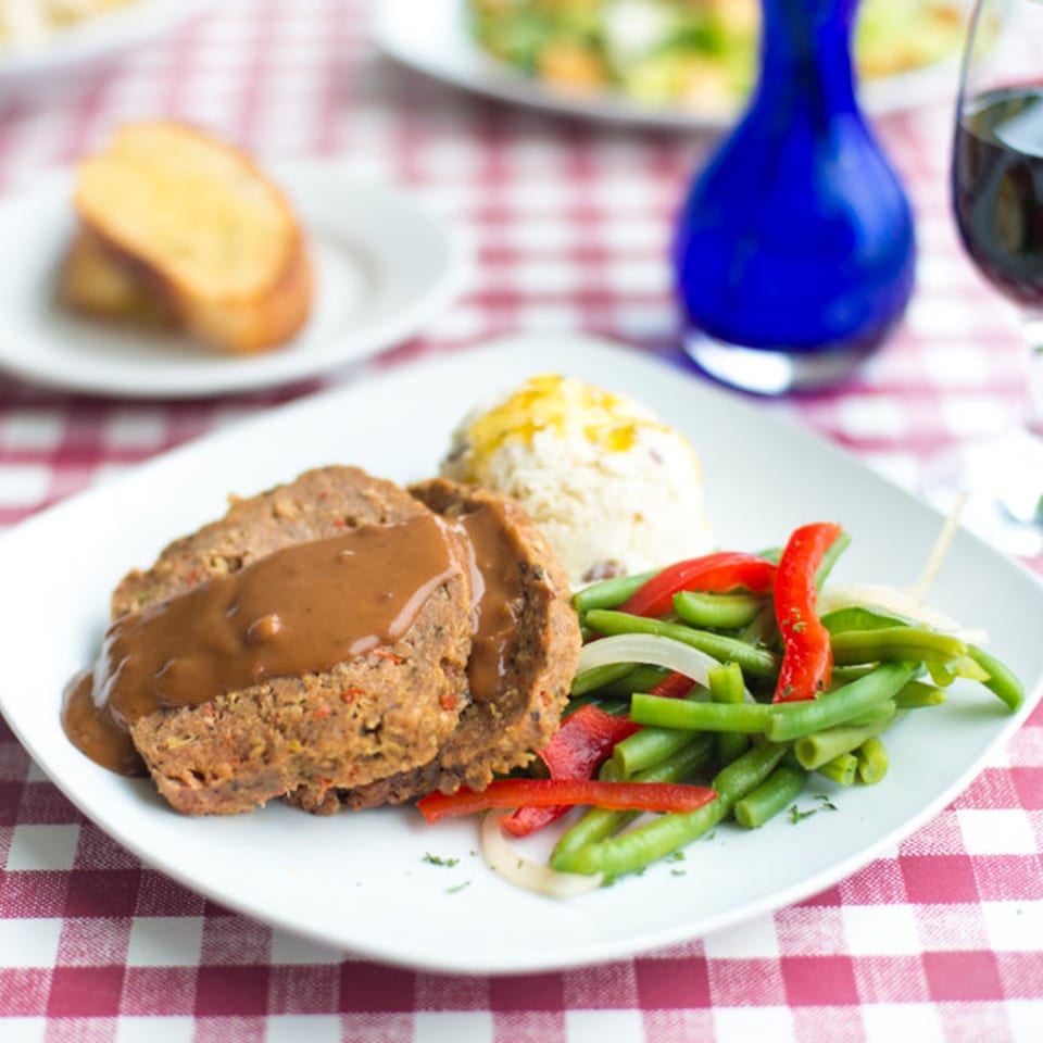 Meatloaf with gravy, mashed potatoes, and green beans with peppers on a red and white checkered tablecloth.