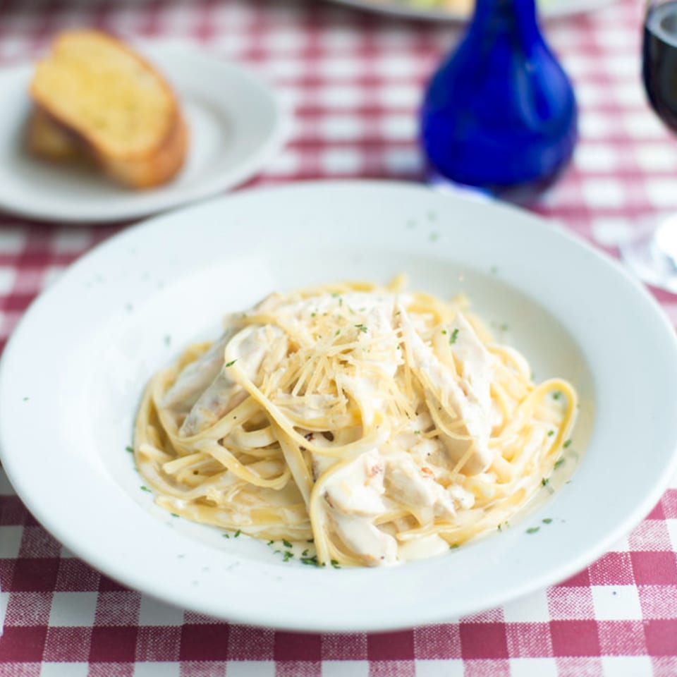 Creamy Alfredo pasta with grated cheese and parsley, served with garlic bread on a red checkered tablecloth.